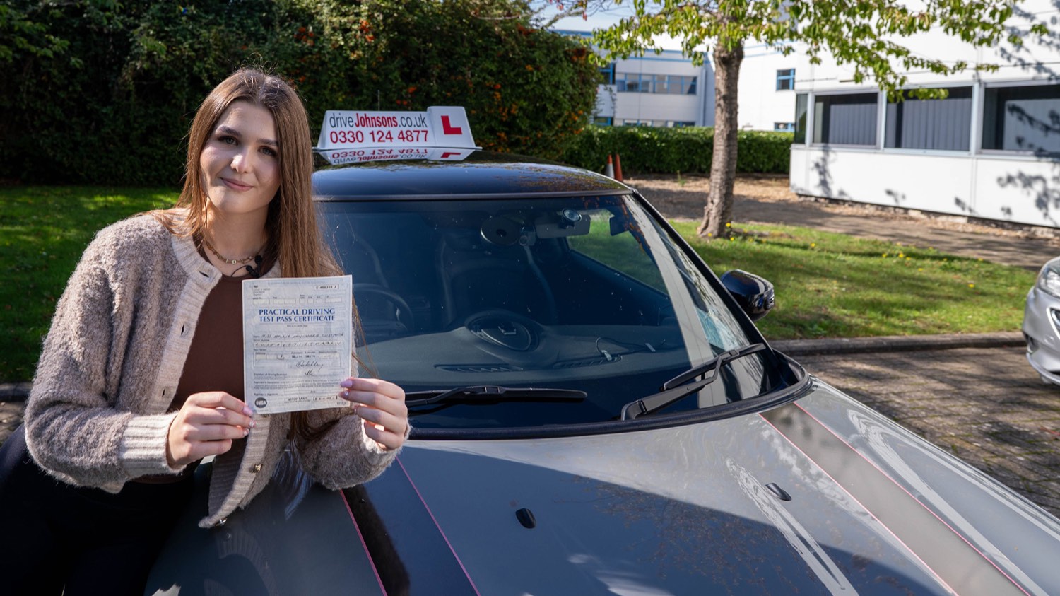 A driveJohnson’s learner holding her driving test pass certificate beside a driving school car