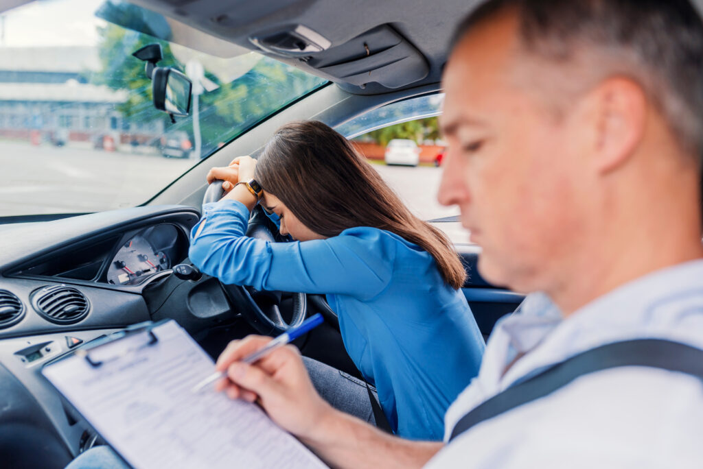 Learner who has failed their driving test with their head on the steering wheel next to driving examiner